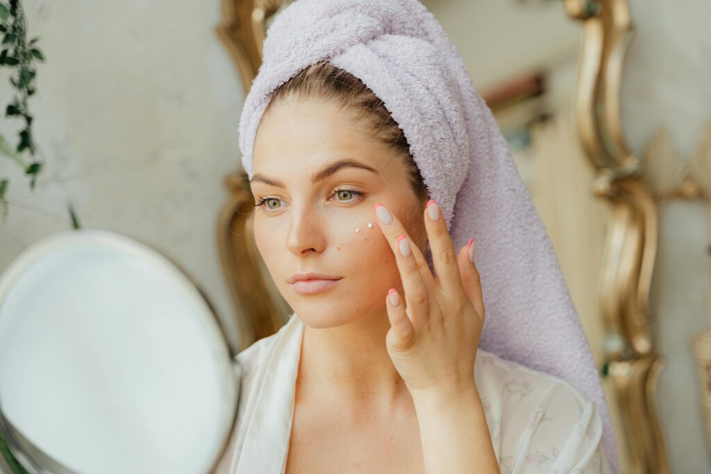 pexels-photo-5205669-5205669 Woman with head towel applying moisturizer in front of mirror indoors.